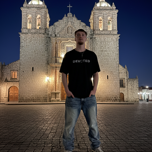 Person wearing a black t-shirt with 'DEVOTED' text in front of a large stone building at night.
